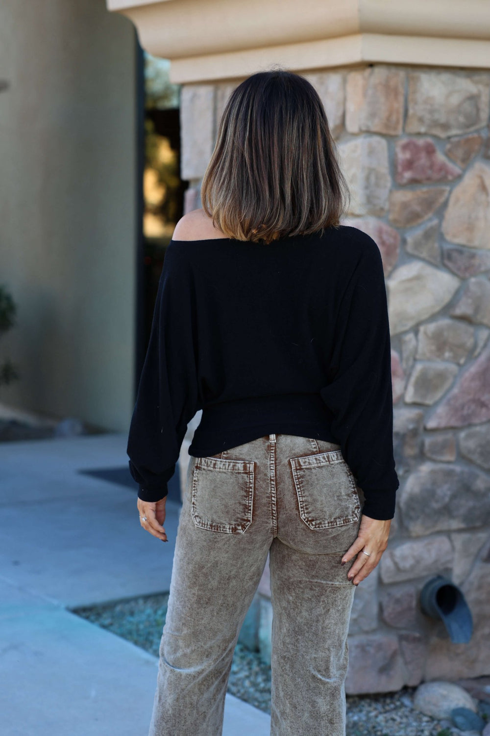 A woman in a Black Off The Shoulder Crepe Top and beige jeans stands outdoors with her shoulder-length hair flowing, facing away.