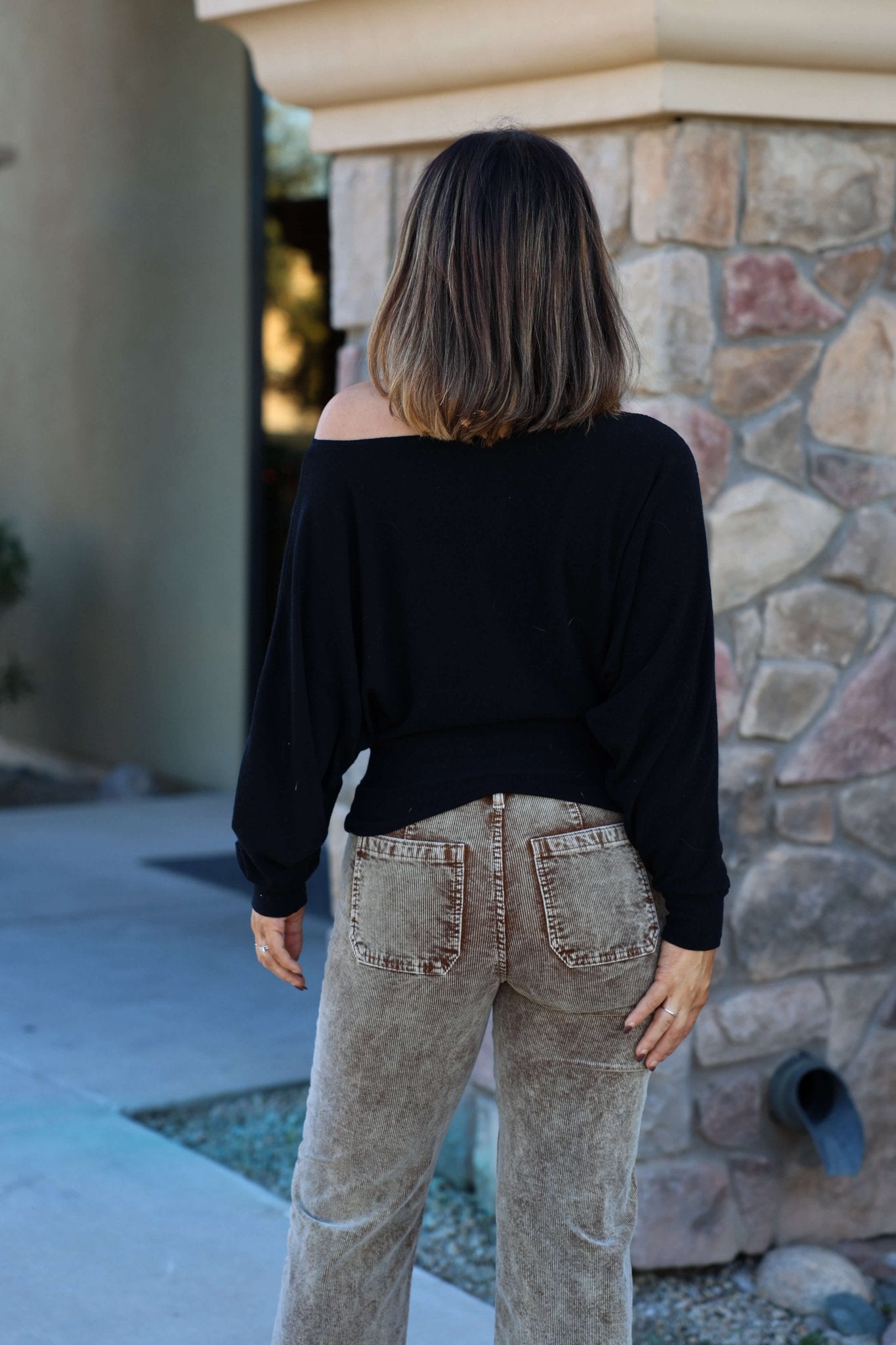 A woman in a Black Off The Shoulder Crepe Top and beige jeans stands outdoors with her shoulder-length hair flowing, facing away.