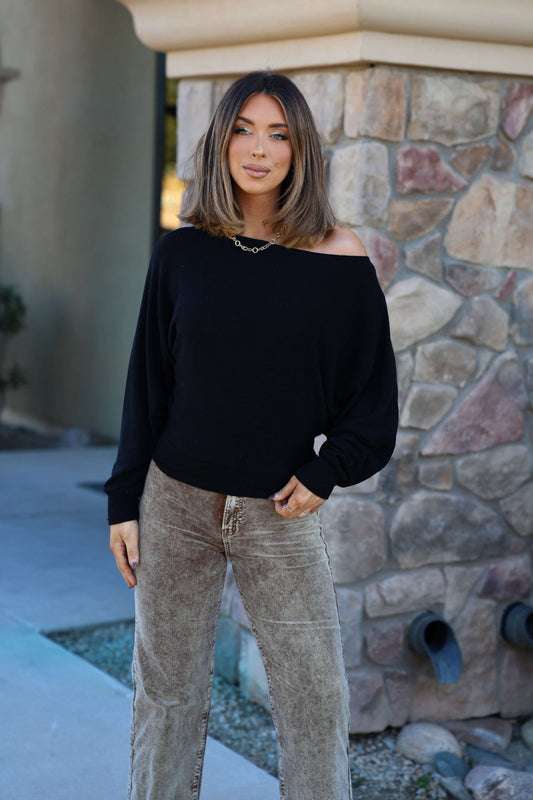A woman in a Black Off The Shoulder Crepe Top and light brown pants stands by a stone wall outdoors, her hair styled neatly.