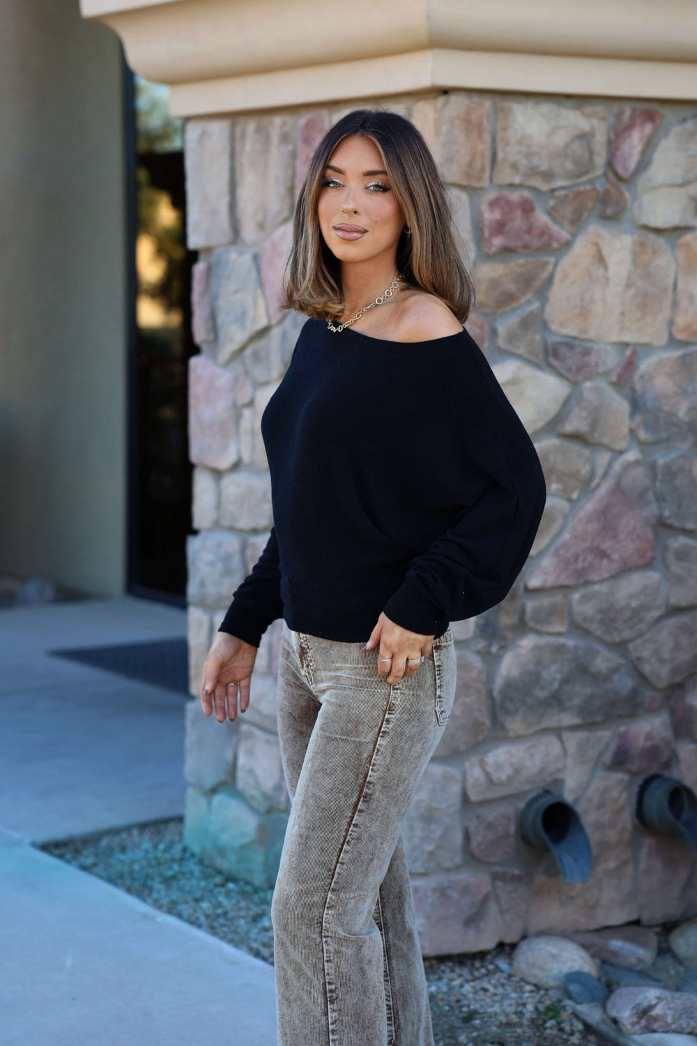 A woman with shoulder-length hair wears a Black Off The Shoulder Crepe Top and light-colored jeans in front of a stone wall.
