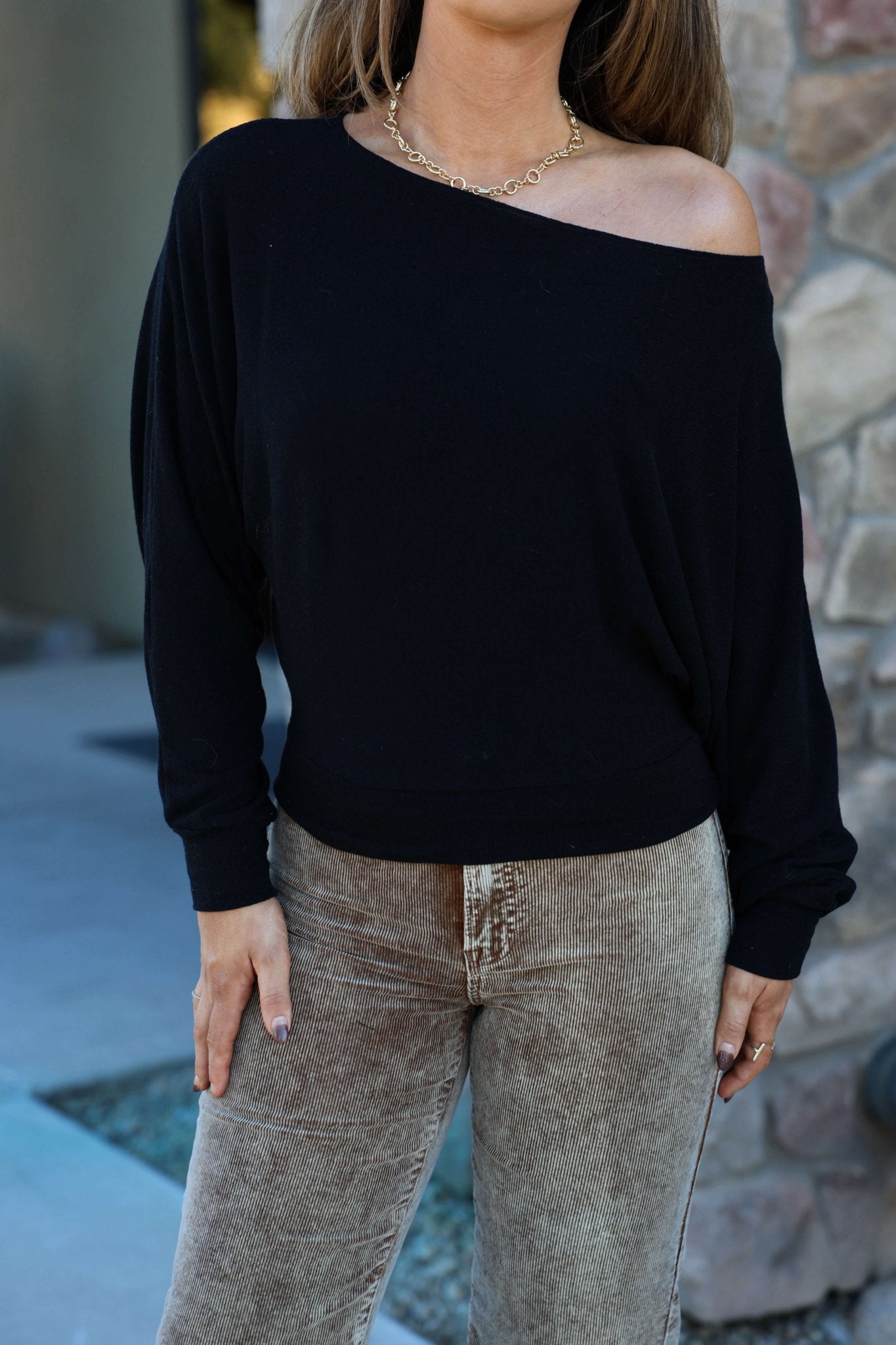 A woman in a Black Off The Shoulder Crepe Top, beige corduroy pants, and a gold chain necklace stands by a stone wall.