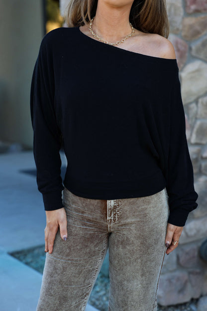 A woman in a Black Off The Shoulder Crepe Top, beige corduroy pants, and a gold chain necklace stands by a stone wall.