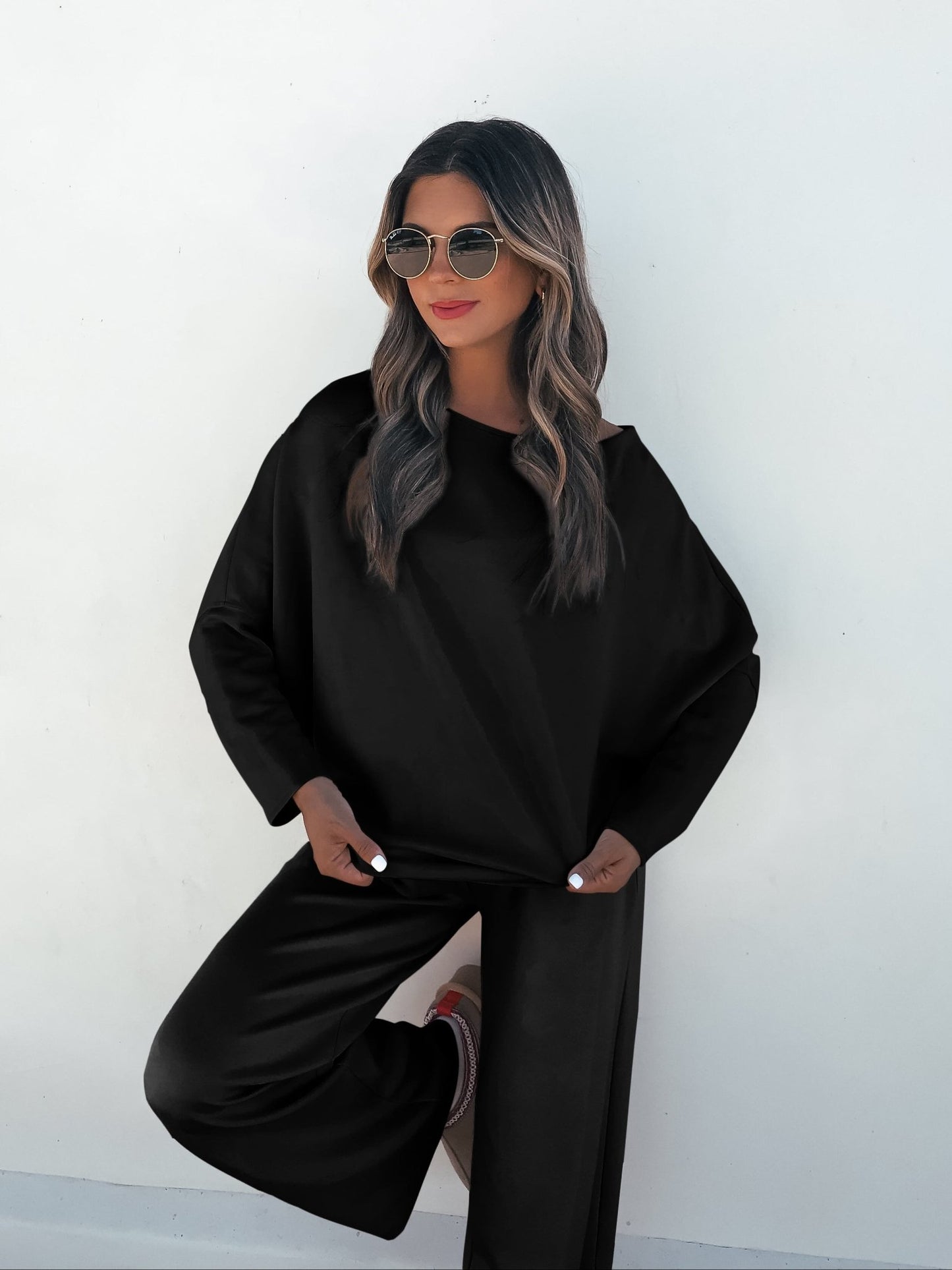 A woman confidently poses in a Black Off The Shoulder Scuba Top and sunglasses against a white wall.