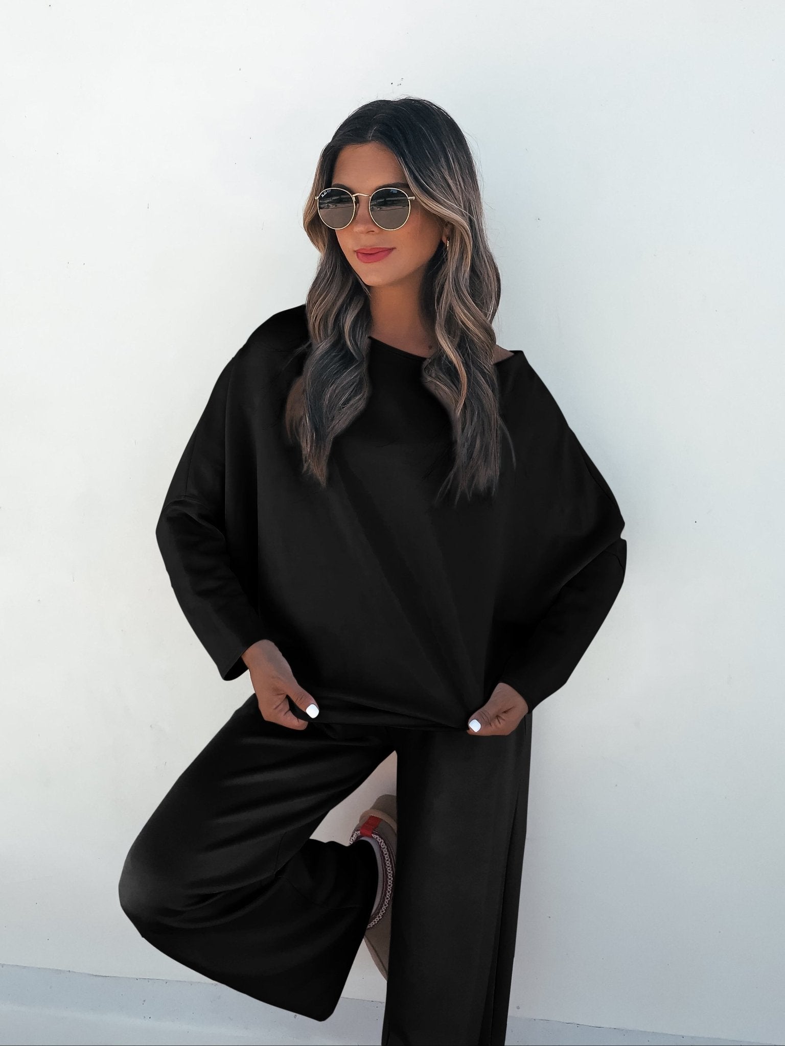 A woman confidently poses in a Black Off The Shoulder Scuba Top and sunglasses against a white wall.
