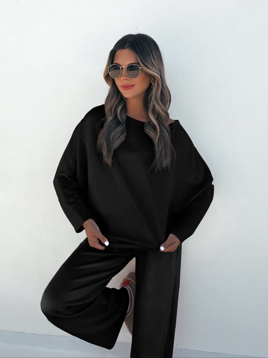 A woman confidently poses in a Black Off The Shoulder Scuba Top and sunglasses against a white wall.