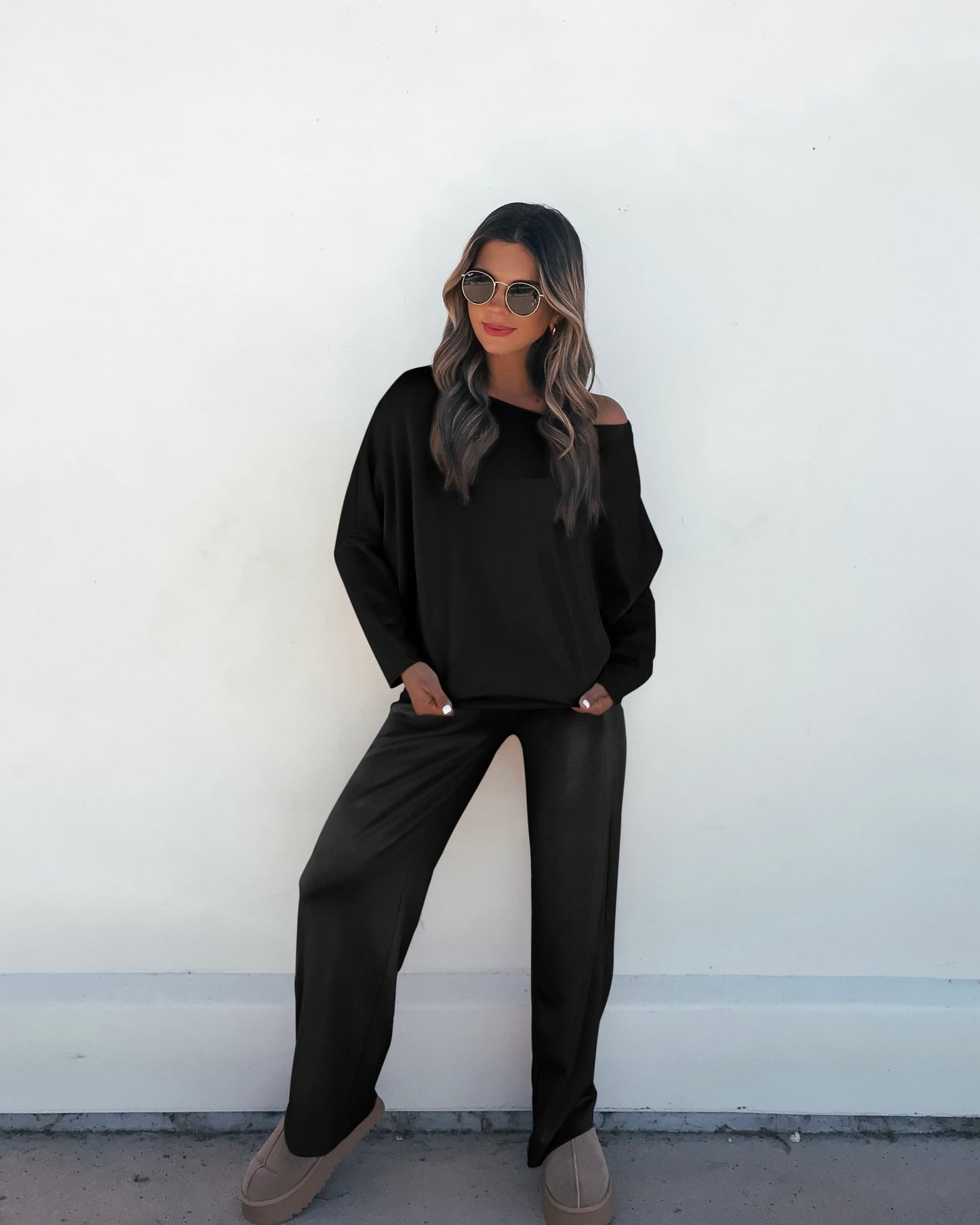 A woman in a Black Off The Shoulder Scuba Top stands relaxed, hands in pockets, against a plain white wall.