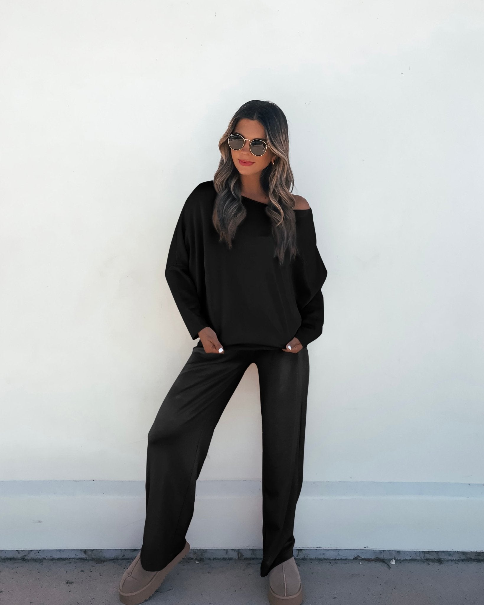 A woman in a Black Off The Shoulder Scuba Top stands relaxed, hands in pockets, against a plain white wall.