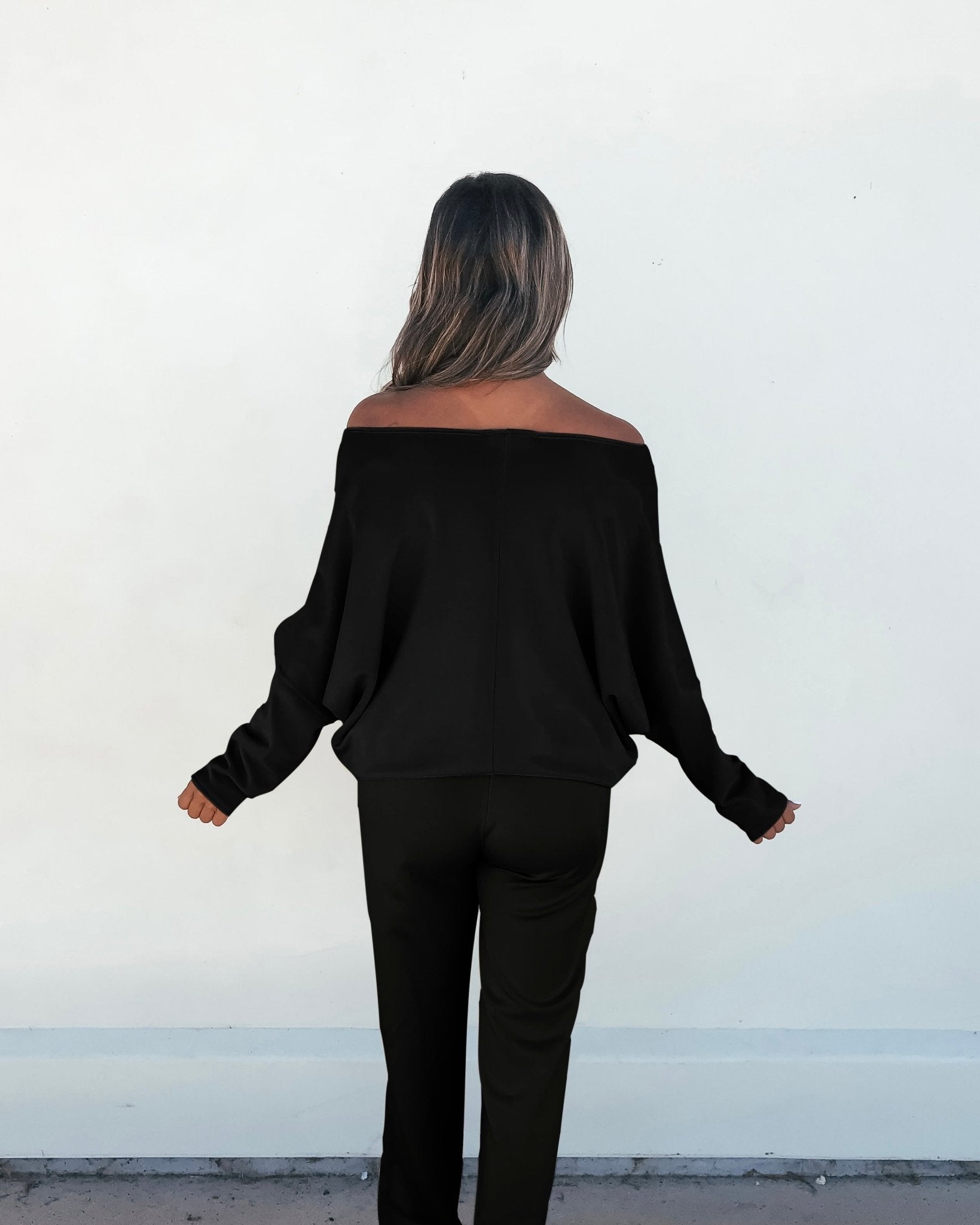 A person with long hair faces a white wall, wearing the Black Off The Shoulder Scuba Top and black pants.