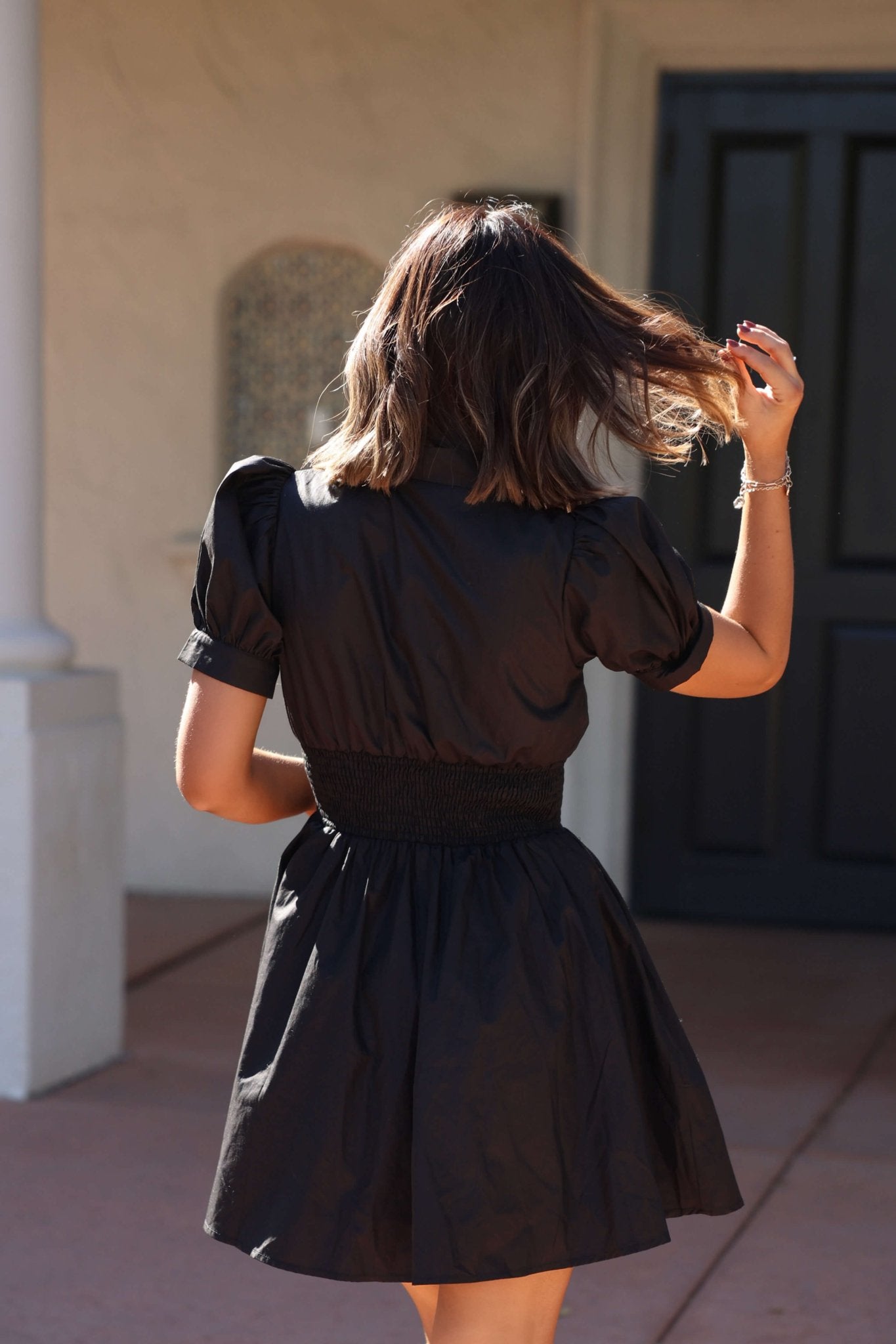 A woman in a Black Puff Sleeve Smocked Mini Dress - FINAL SALE stands outside, facing away, touching her hair.
