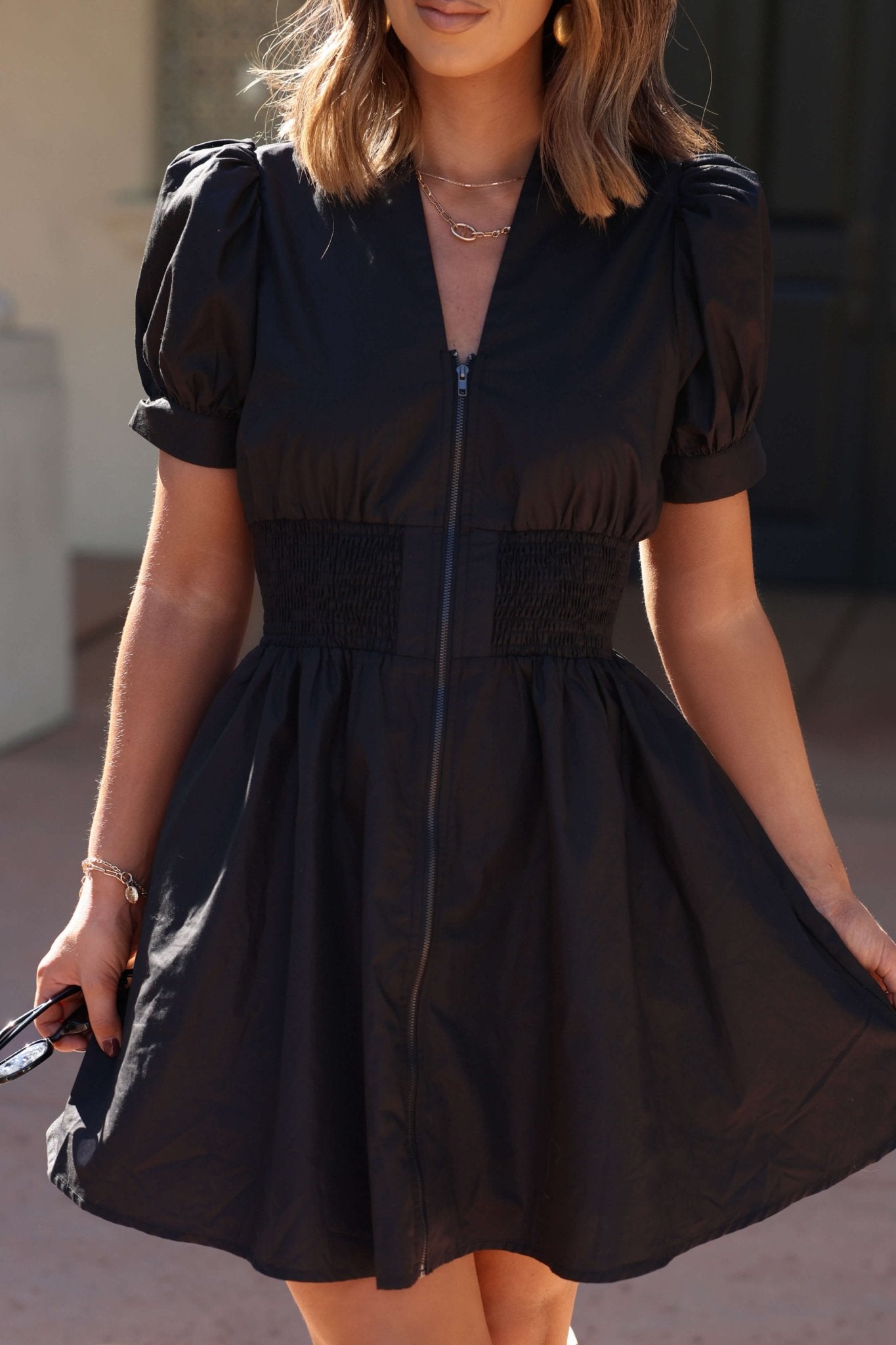 Woman wearing the Black Puff Sleeve Smocked Mini Dress - FINAL SALE, holding sunglasses in one hand.
