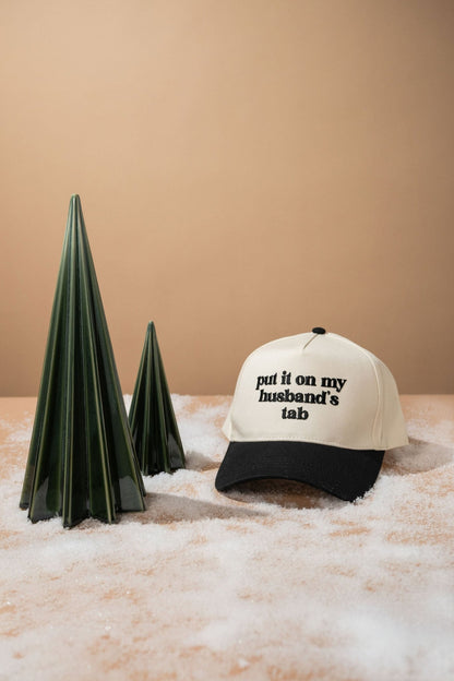 The Black Put It On My Husband's Tab Baseball Cap is displayed on fake snow next to two green cone-shaped trees.