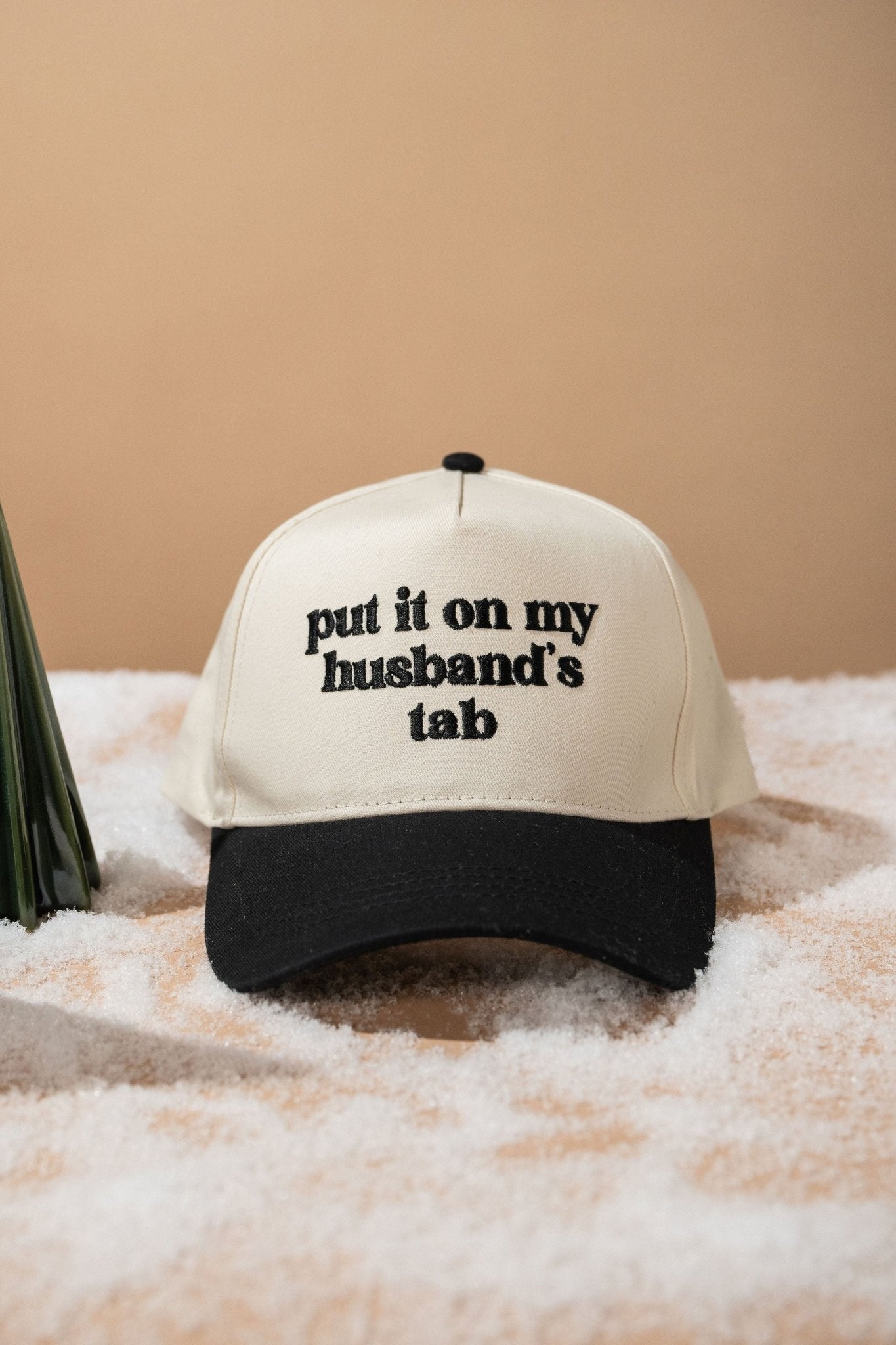 The Black Put It On My Husband's Tab Baseball Cap sits on a snowy surface, featuring adjustable cotton and embroidered text.
