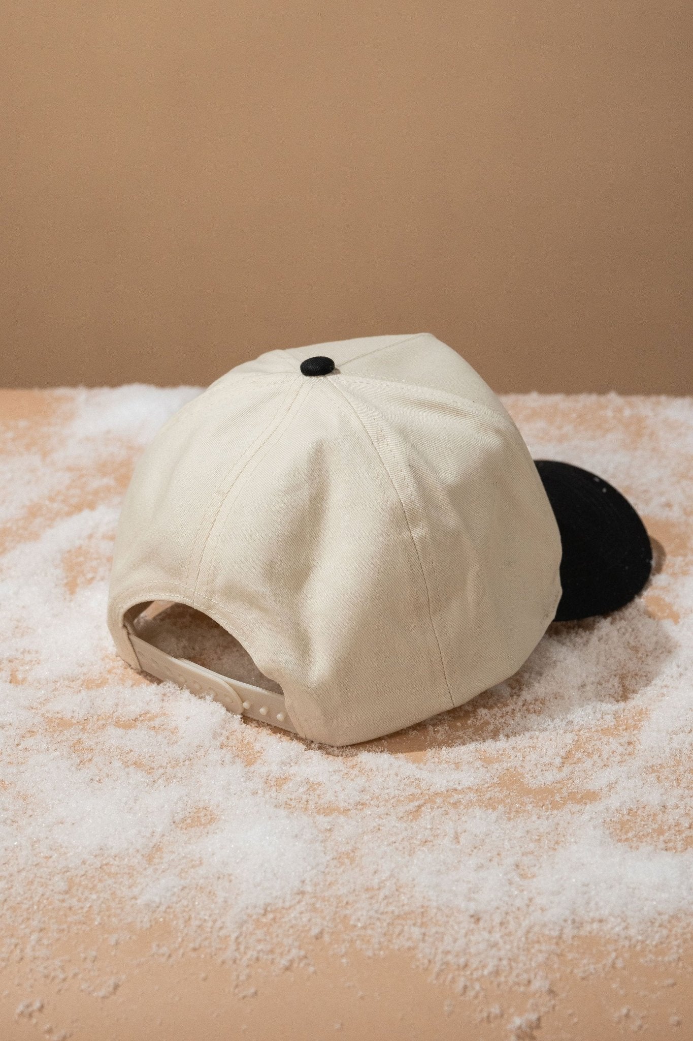 A Black Put It On My Husband's Tab Baseball Cap rests upside down on a tan, powder-dusted surface with a beige background.