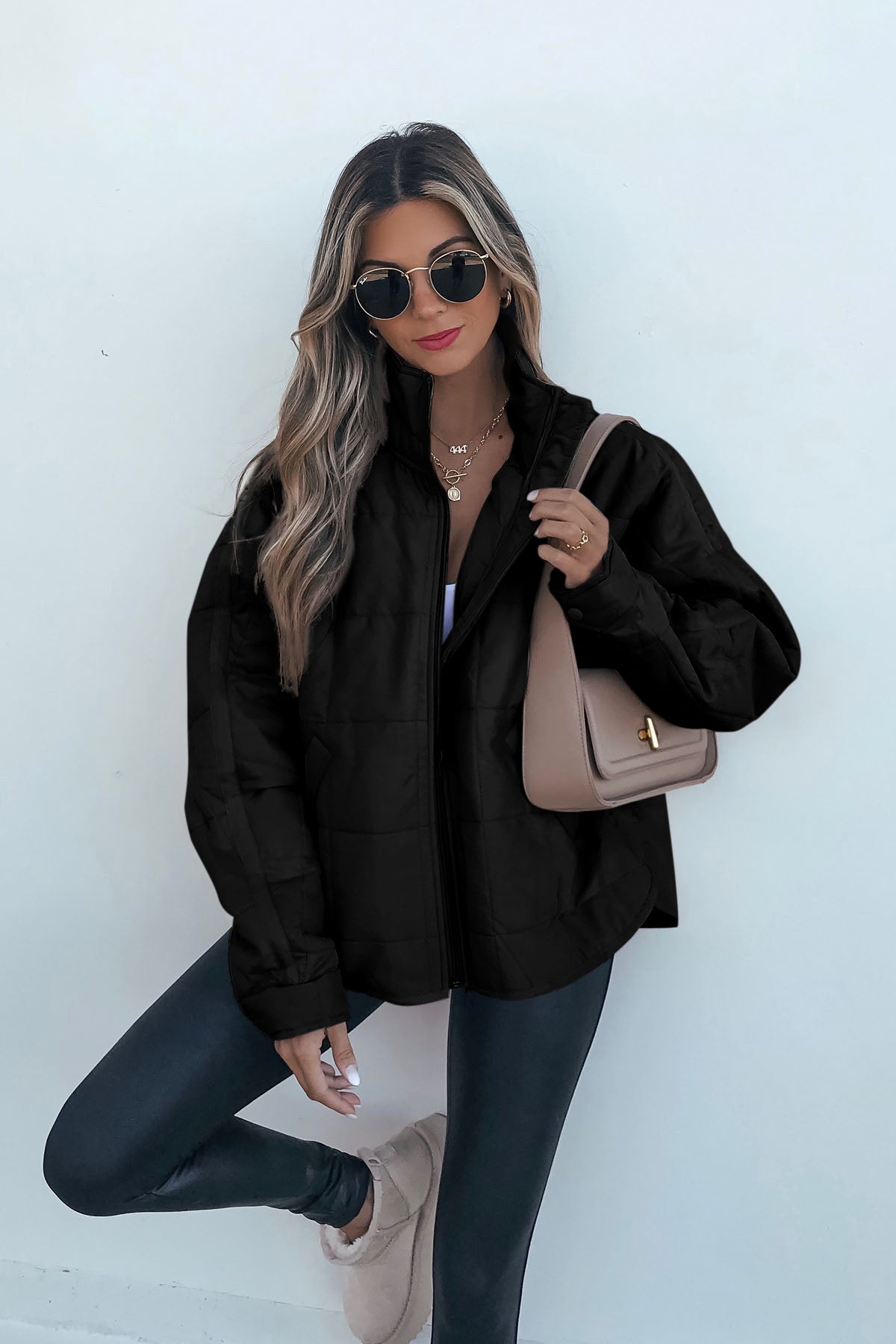 Woman with long blonde hair wears sunglasses, black quilted zip up puffer jacket, black leggings, and beige purse against a white background.