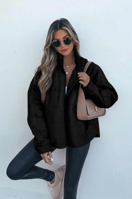 Woman with long blonde hair wears sunglasses, black quilted zip up puffer jacket, black leggings, and beige purse against a white background.