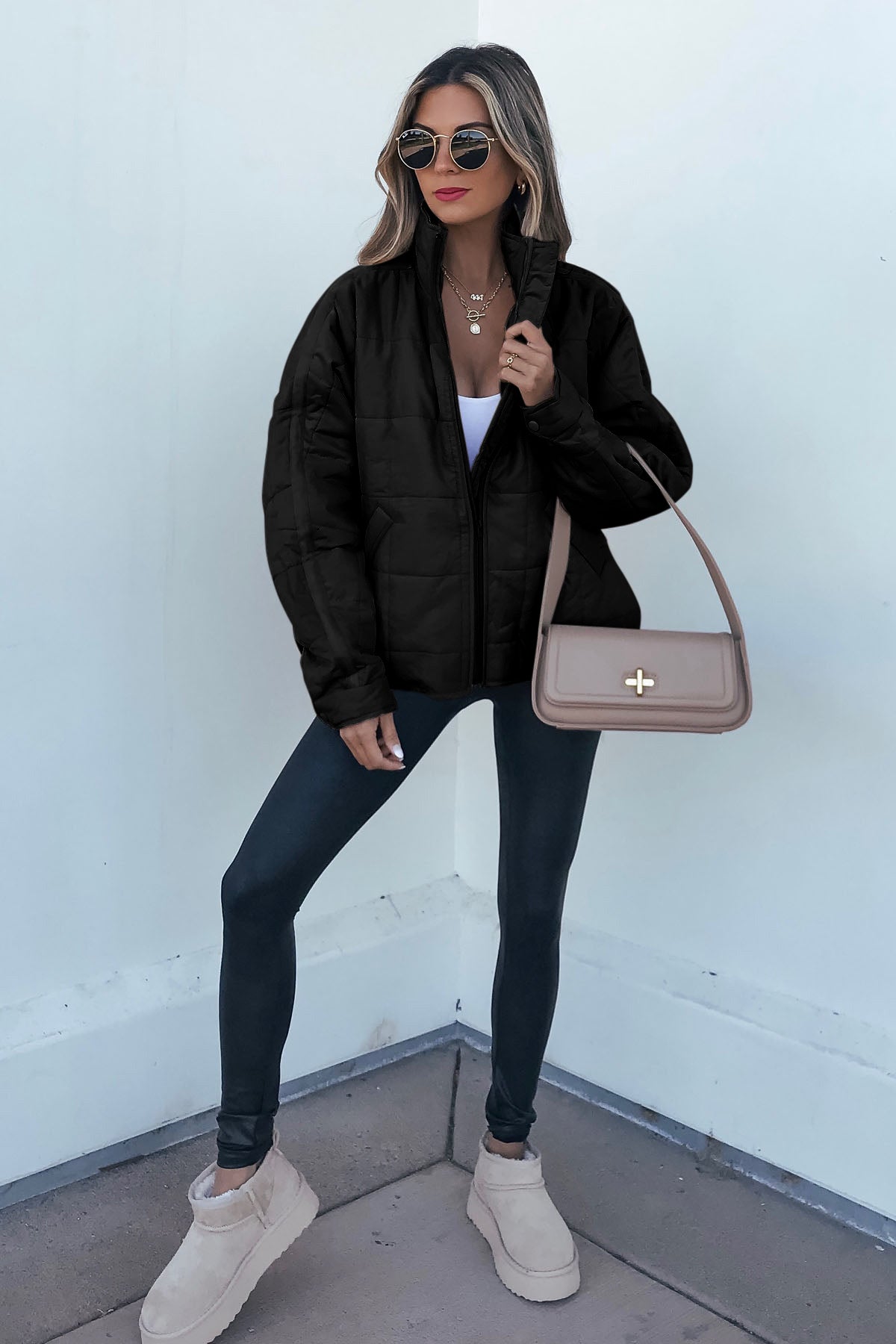 A woman in sunglasses wears a Black Quilted Zip Up Puffer Jacket, black leggings, beige boots, and a beige shoulder bag by a white wall.