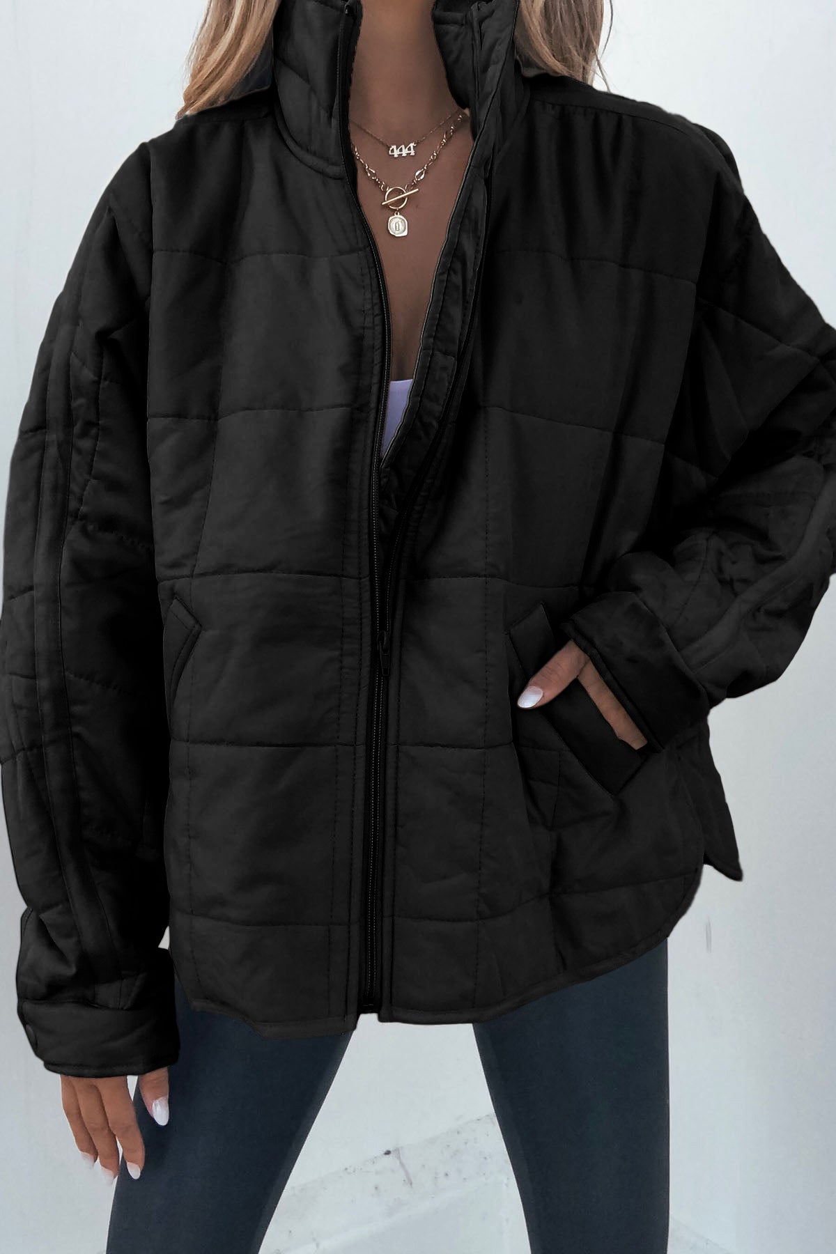 A person wears the Black Quilted Zip Up Puffer Jacket, dark leggings, layered gold necklaces, and has a hand in their pocket.