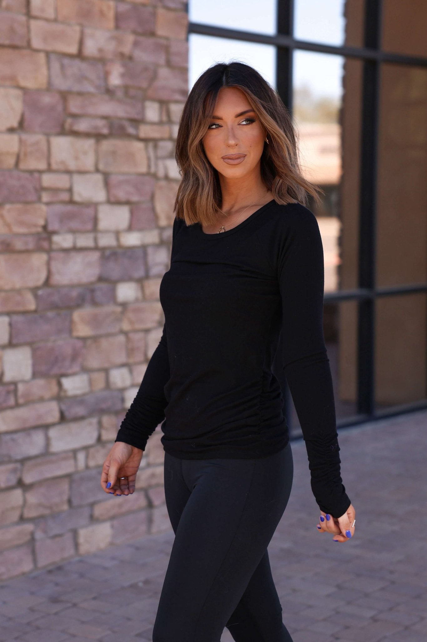 Woman with shoulder-length brown hair in a Black Ruched Sleeve Ribbed Top - DOORBUSTER and black leggings walks by a stone wall.