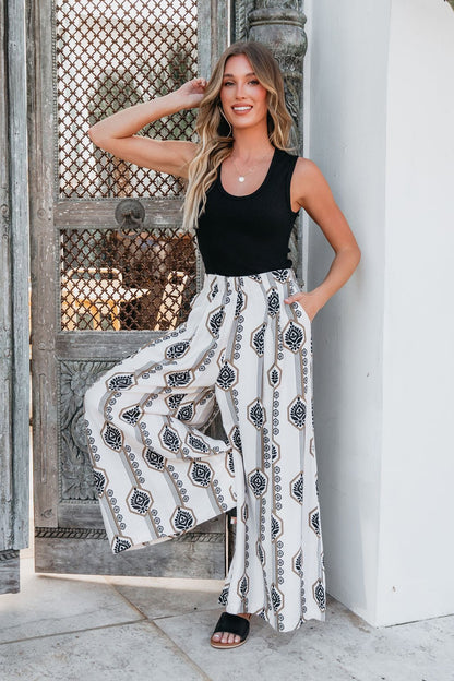 Woman in a Black Scoop Neck Ribbed Tank Top smiles by an ornate wooden door, one arm raised and the other in her pocket.