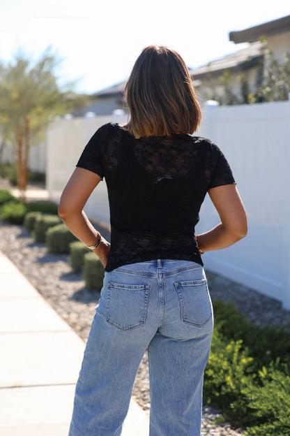 A woman faces away from the camera in a Black Sheer Floral Lace Top and light blue jeans on a sunny day.