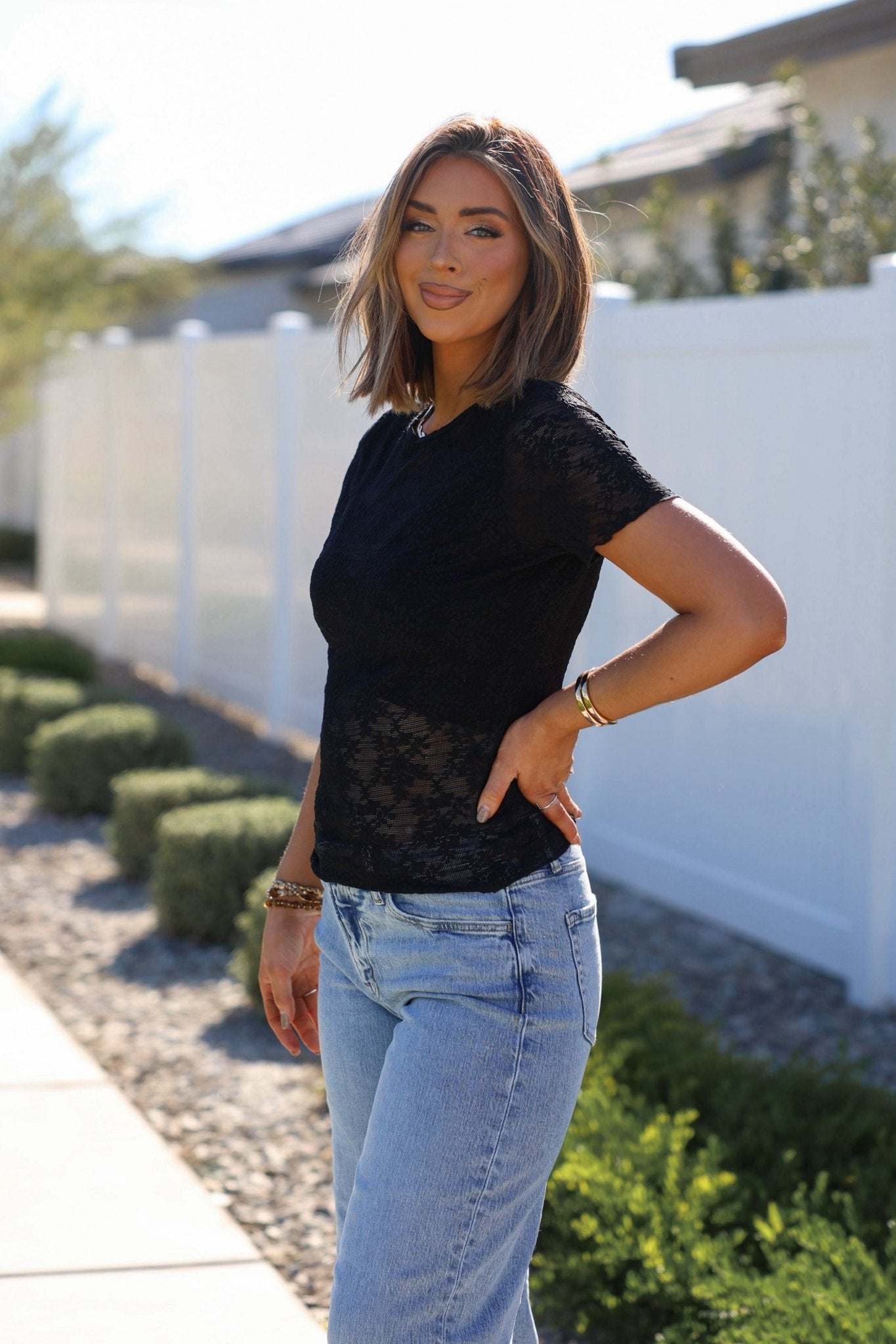 Smiling outdoors by a white fence, a woman in the Black Sheer Floral Lace Top and light blue jeans shows off her style.