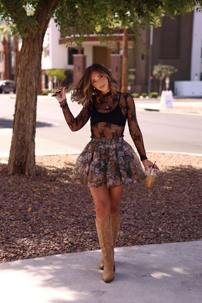 Woman in a Black Sheer Lace Mock Neck Top poses outside on a sunny day, holding iced coffee and wearing tan boots.
