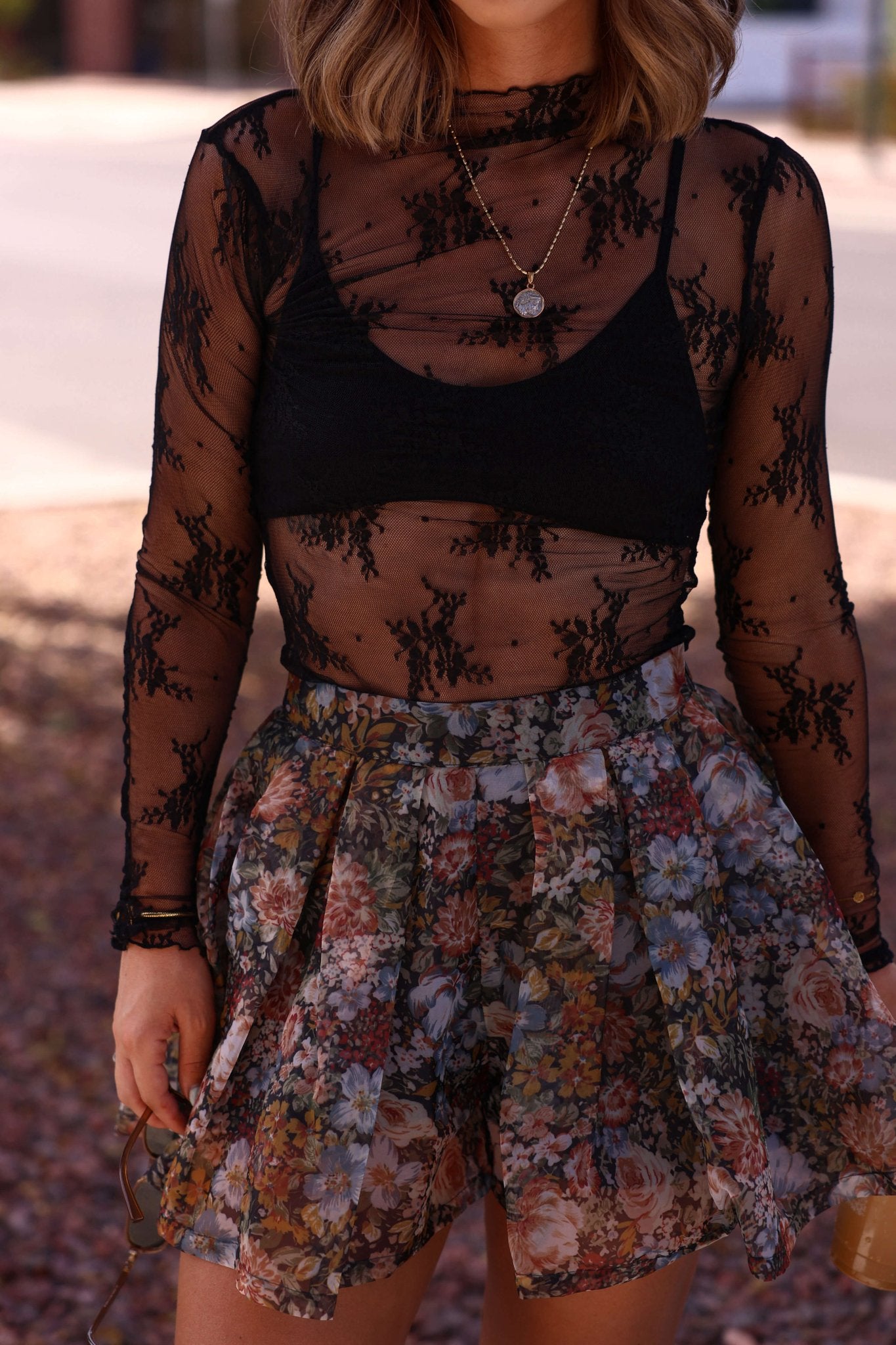 Woman wears the Black Sheer Lace Mock Neck Top - DOORBUSTER over a black bra, paired with a high-waisted floral skirt.