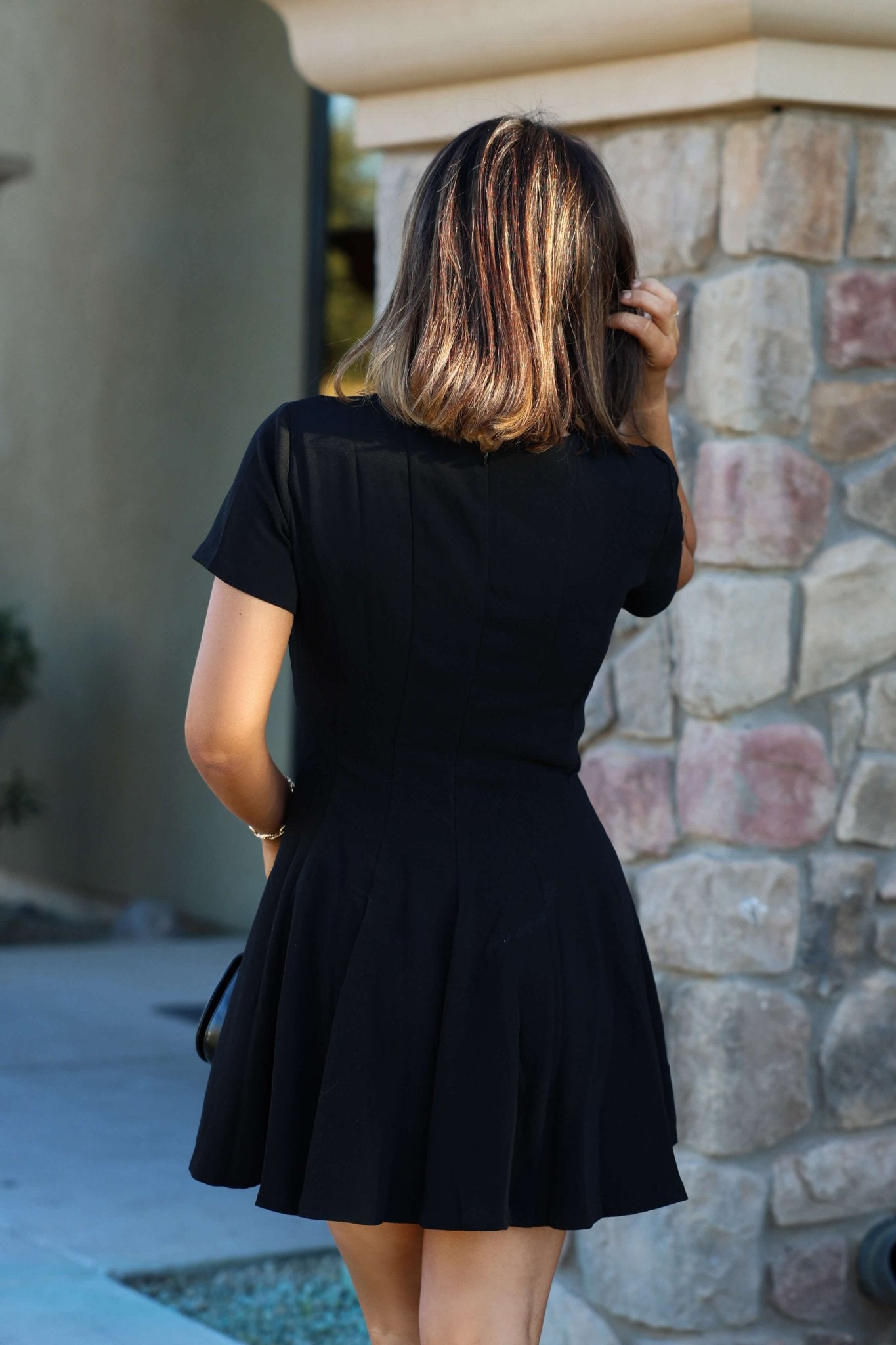 A woman with shoulder-length hair stands outdoors in a Black Short Sleeve Flare Mini Dress, her back to the camera by a stone wall.