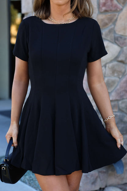 Woman wearing a Black Short Sleeve Flare Mini Dress, gold jewelry, and holding a black handbag in front of a stone wall.