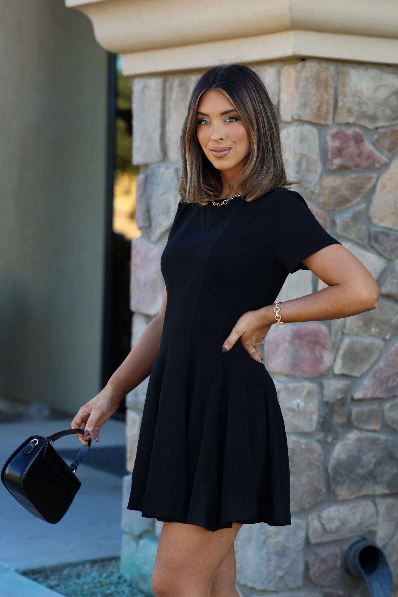 Woman wearing a Black Short Sleeve Flare Mini Dress, holding a small purse and smiling by a stone wall.