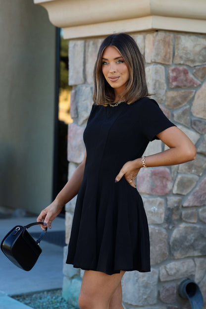 Woman wearing a Black Short Sleeve Flare Mini Dress, holding a small purse and smiling by a stone wall.