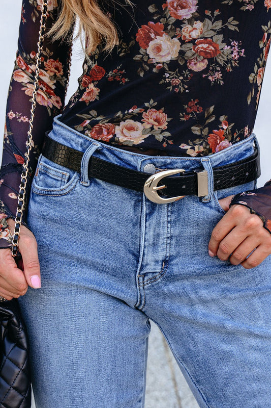Woman in high-waisted blue jeans, black floral top, a black snake skin leather belt, and holding a quilted black purse.