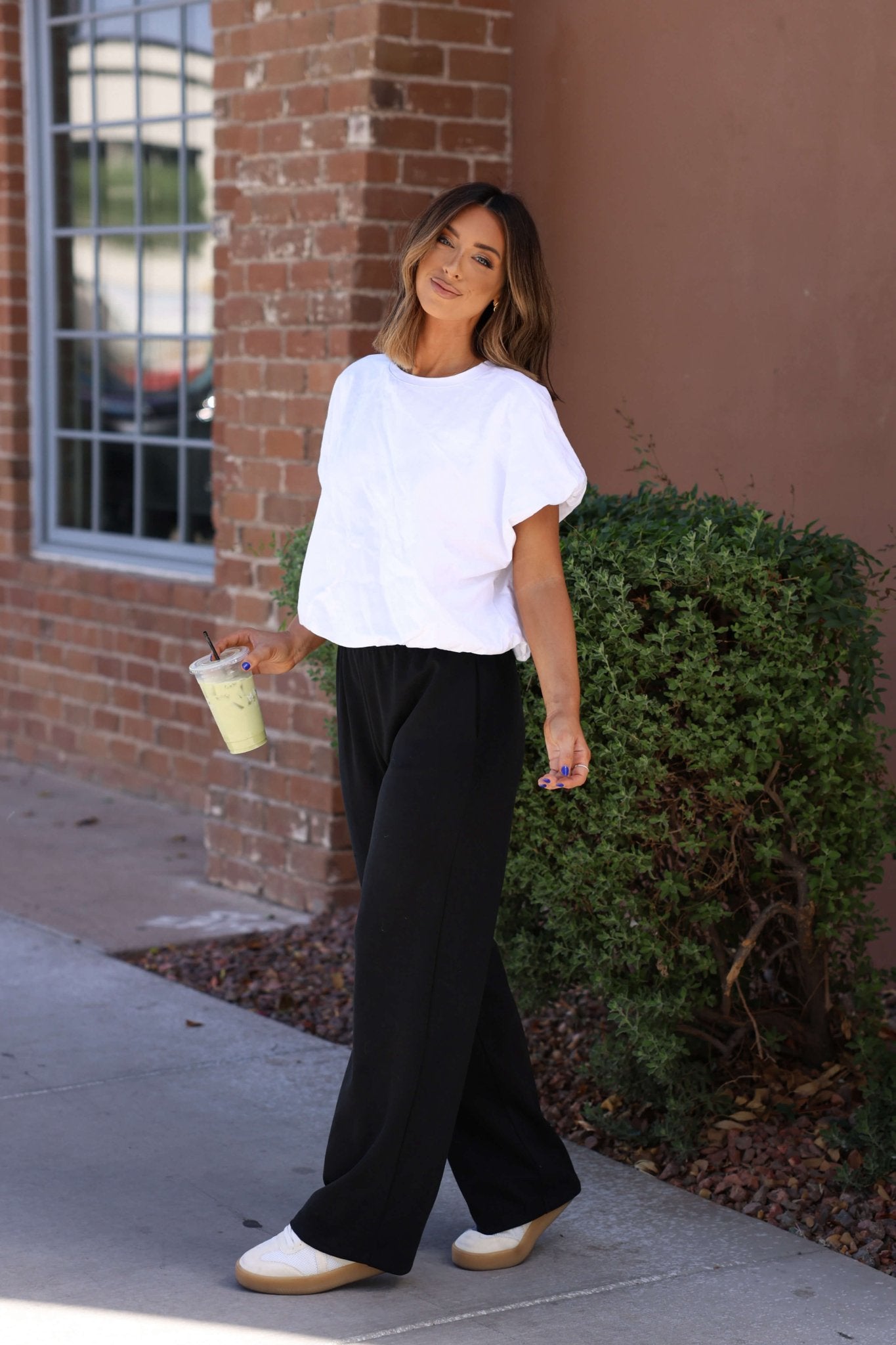 Woman in a white t-shirt and Black Soft Cloud Scuba Sweatpants stands outside by a brick wall, holding a drink and smiling.