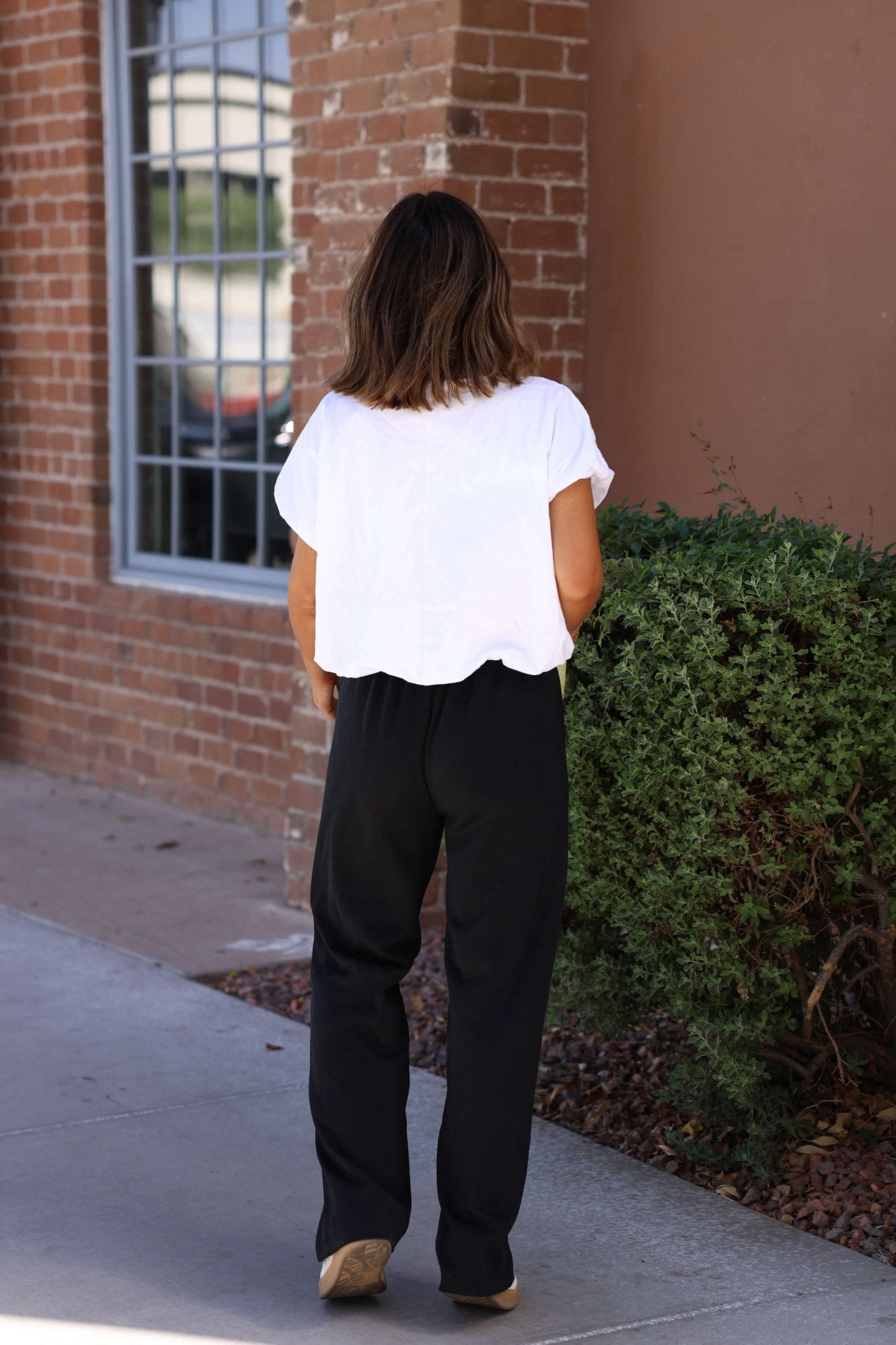 Wearing Black Soft Cloud Scuba Sweatpants and a white top, a person stands outdoors by a brick wall and green bush.