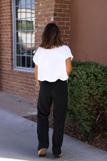 Wearing Black Soft Cloud Scuba Sweatpants and a white top, a person stands outdoors by a brick wall and green bush.