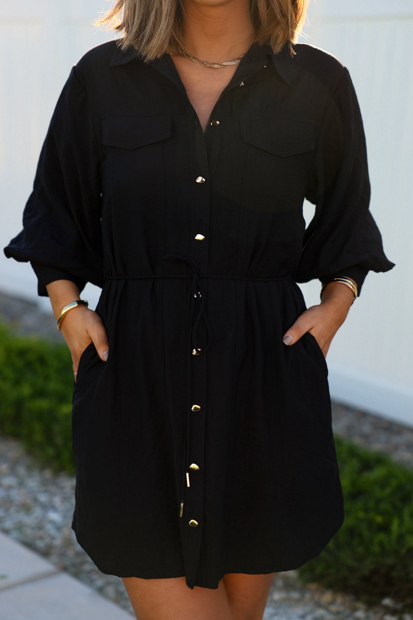 Wearing the Black Tie Mini Shirt Dress with adjustable waist, a woman stands outdoors, hands in pockets, gold bracelets and necklace on.