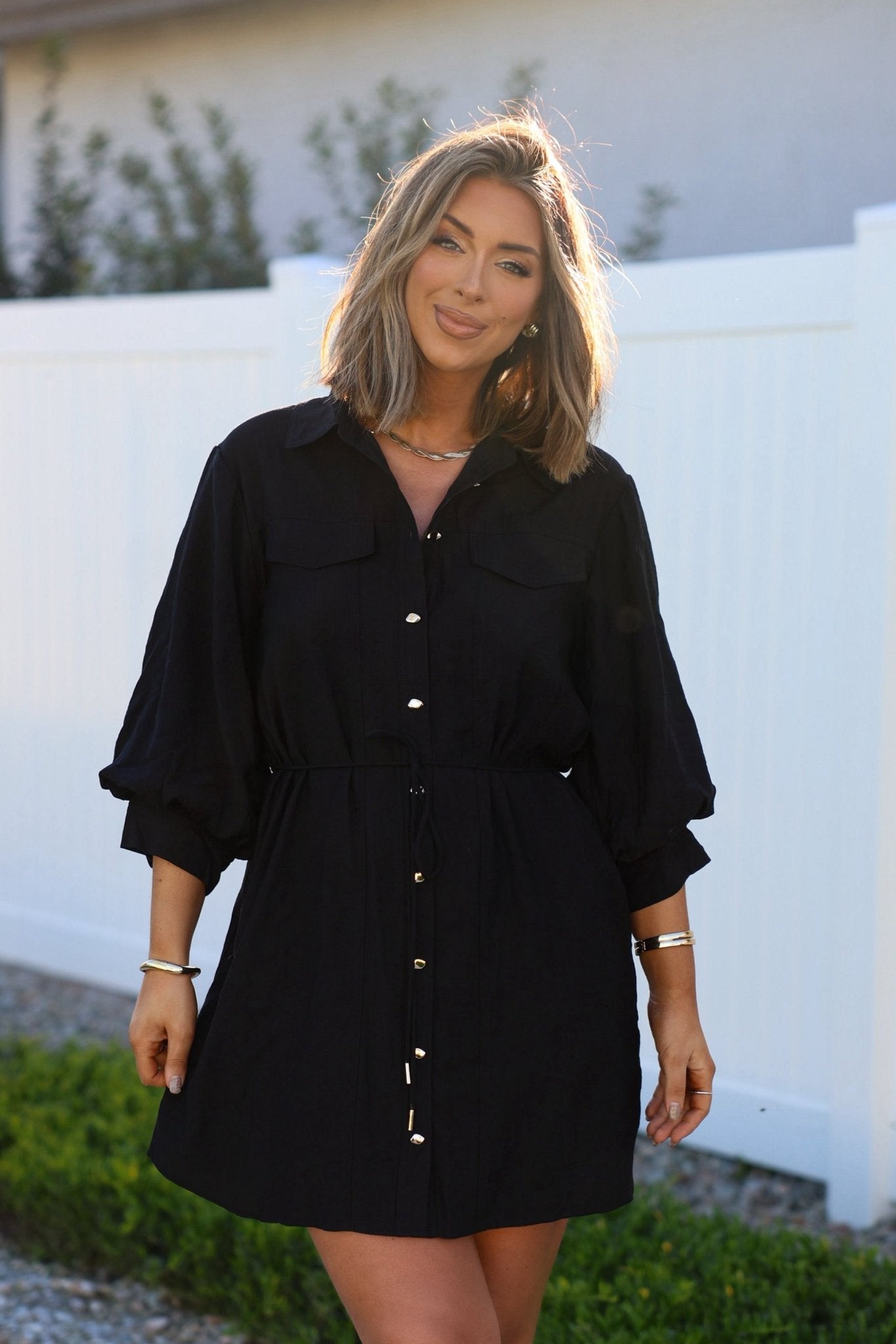 Smiling woman outdoors by a white fence wears the Black Tie Mini Shirt Dress with adjustable waist.
