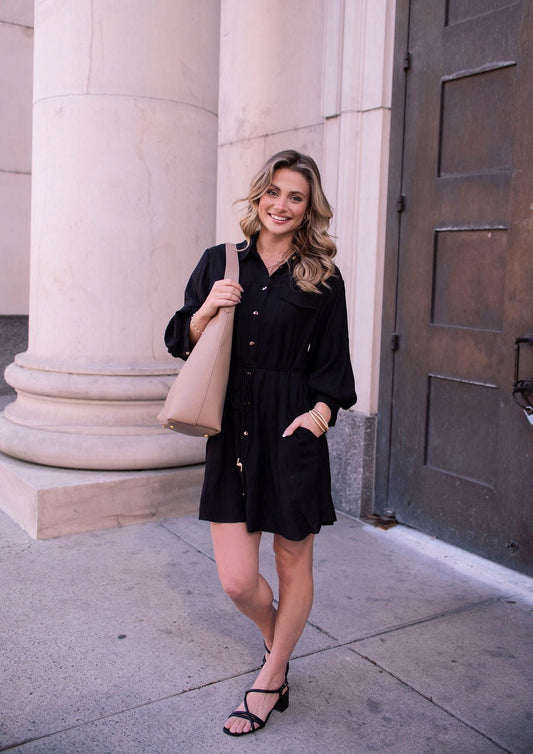 Woman in a Black Tie Mini Shirt Dress and sandals stands by a column, smiling and holding a beige shoulder bag.