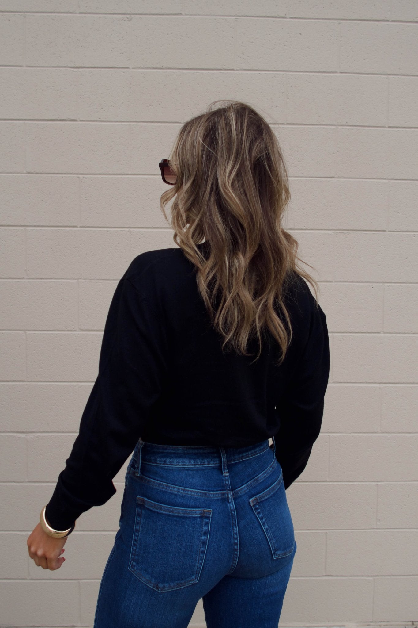 A woman with wavy hair in sunglasses and blue jeans wears a Black V Neck Buttoned Cardigan, standing by a beige brick wall.