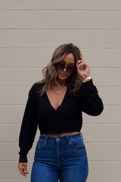 Woman in a Black V Neck Buttoned Cardigan and sunglasses poses by a beige brick wall, holding her sunglasses.