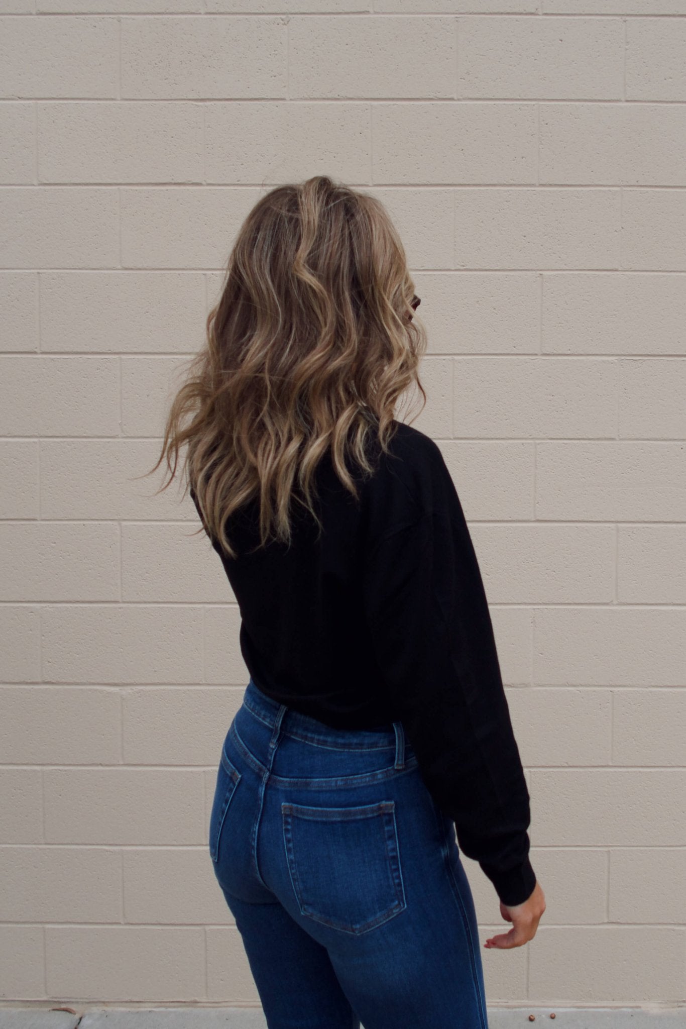 Woman with wavy blonde hair wears a Black V Neck Buttoned Cardigan and blue jeans, facing a beige brick wall with her back to camera.