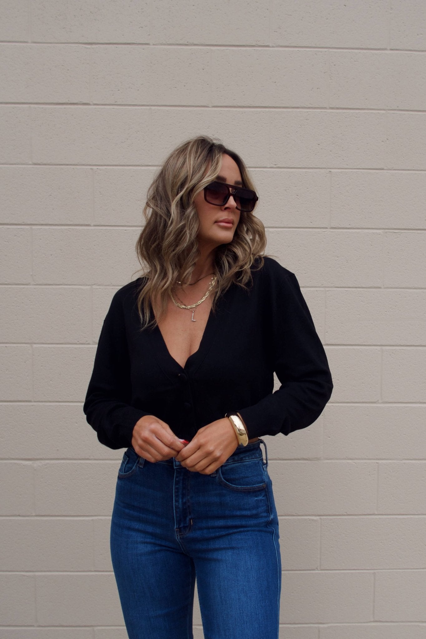Woman with wavy hair in sunglasses and blue jeans wears a Black V Neck Buttoned Cardigan against a beige brick wall.