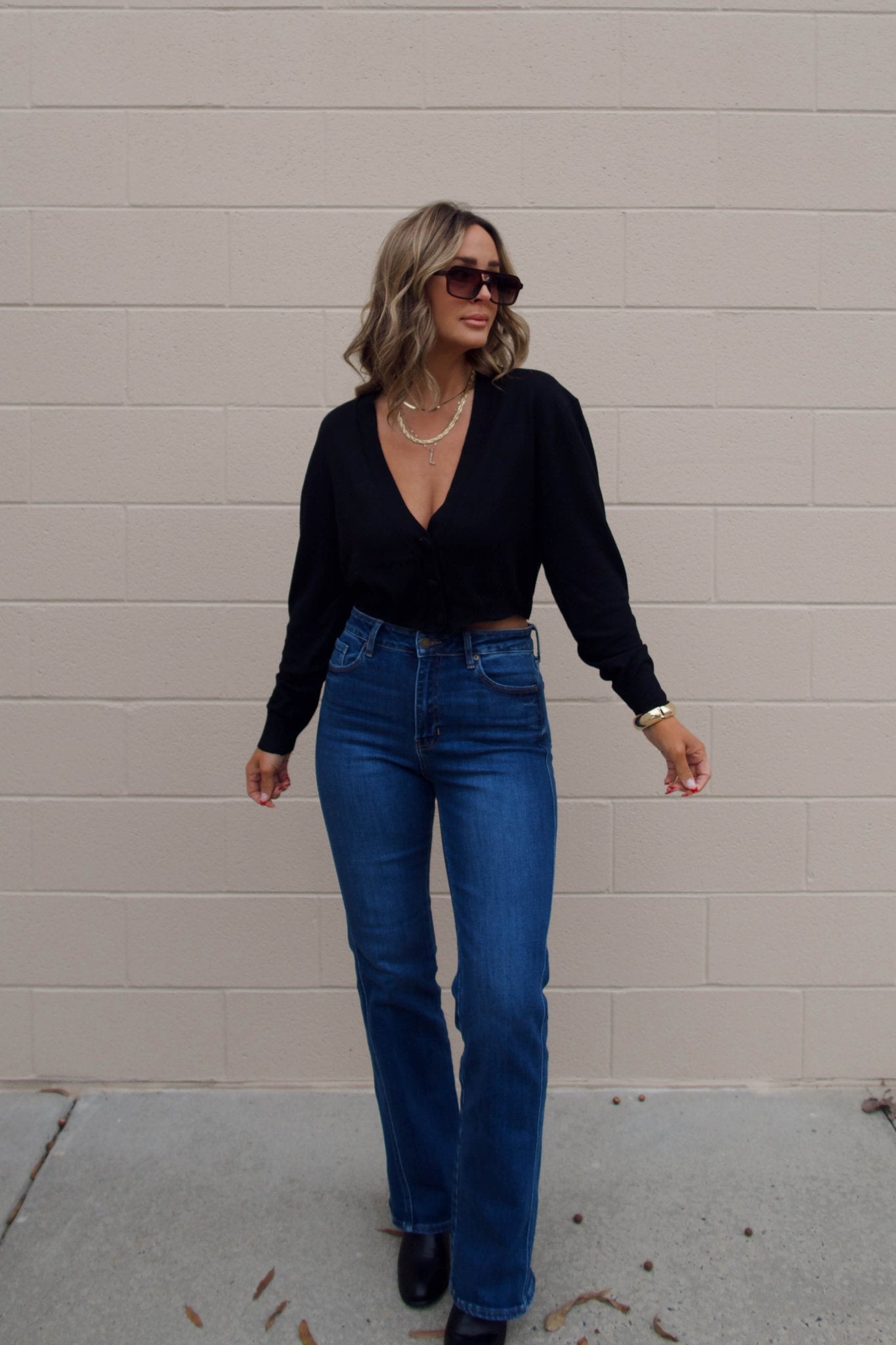 Woman in sunglasses wears a Black V Neck Buttoned Cardigan and blue jeans, smiling with one hand raised against a brick wall.