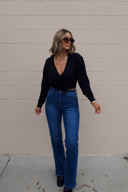 Woman in sunglasses wears a Black V Neck Buttoned Cardigan and blue jeans, smiling with one hand raised against a brick wall.