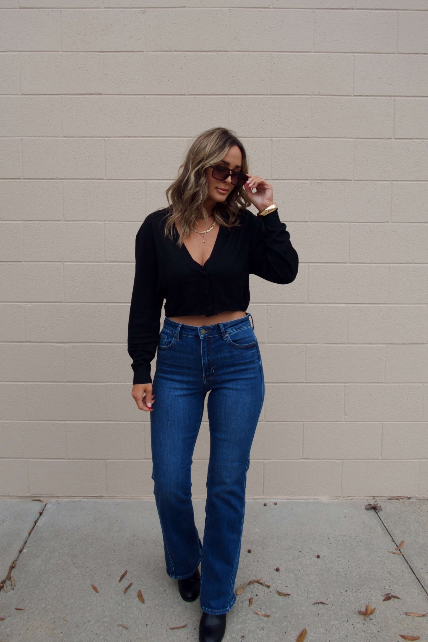 Woman wearing sunglasses and a Black V Neck Buttoned Cardigan with blue jeans stands by a beige brick wall on a concrete sidewalk.