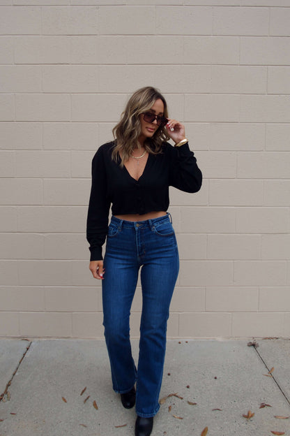 Woman wearing sunglasses and a Black V Neck Buttoned Cardigan with blue jeans stands by a beige brick wall on a concrete sidewalk.