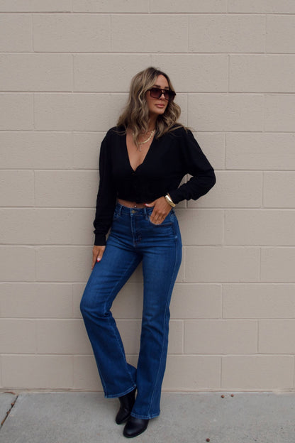 Woman wears a Black V Neck Buttoned Cardigan and blue jeans, standing by a beige brick wall with sunglasses and a slight smile.