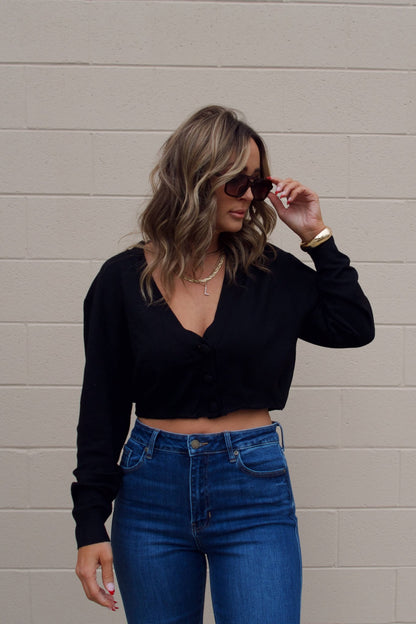 Woman in sunglasses wears a Black V Neck Buttoned Cardigan with high-waisted jeans, standing by a light brick wall.