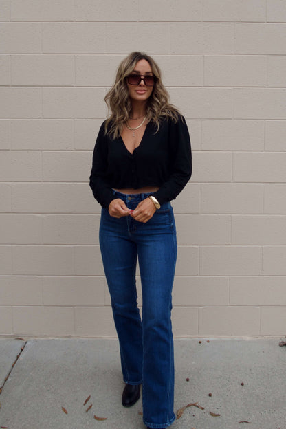 Woman with wavy hair in sunglasses and boots wears the Black V Neck Buttoned Cardigan, blue jeans, by a beige brick wall.