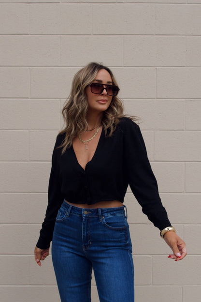 A woman in sunglasses, blue jeans, and a Black V Neck Buttoned Cardigan stands before a beige brick wall.